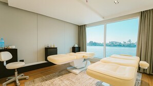 Ein modernes Zimmer mit Massagetisch, einem Sessel, einem kleinen Schrank und einem großen Fenster mit Blick auf ein Gewässer.