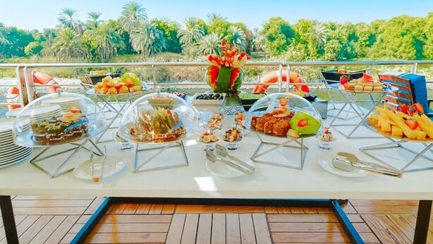 Ein reich gedeckter Buffet mit verschiedenen Speisen und durchsichtigen Abdeckungen, platziert auf einer Terrasse mit Blick auf Palmen und Grünpflanzen.
