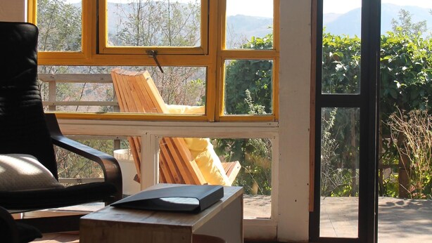 A room with a view of mountains through large windows, a wooden chair, a small table, and a door leading outside. - Sibebe Hills Vista (Mbabane)