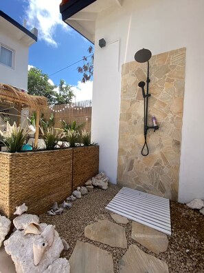 A modern outdoor shower area with a stone wall, a woven planter with plants, and a wooden deck. - Palm Beach Vacaysuites - Coconut Suite - Top Floor (Noord)
