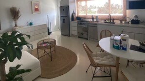 A modern kitchen with white cabinetry, a large window, and a dining area with a round table and chairs. - Palm Beach Vacaysuites - Coconut Suite - Top Floor (Noord)