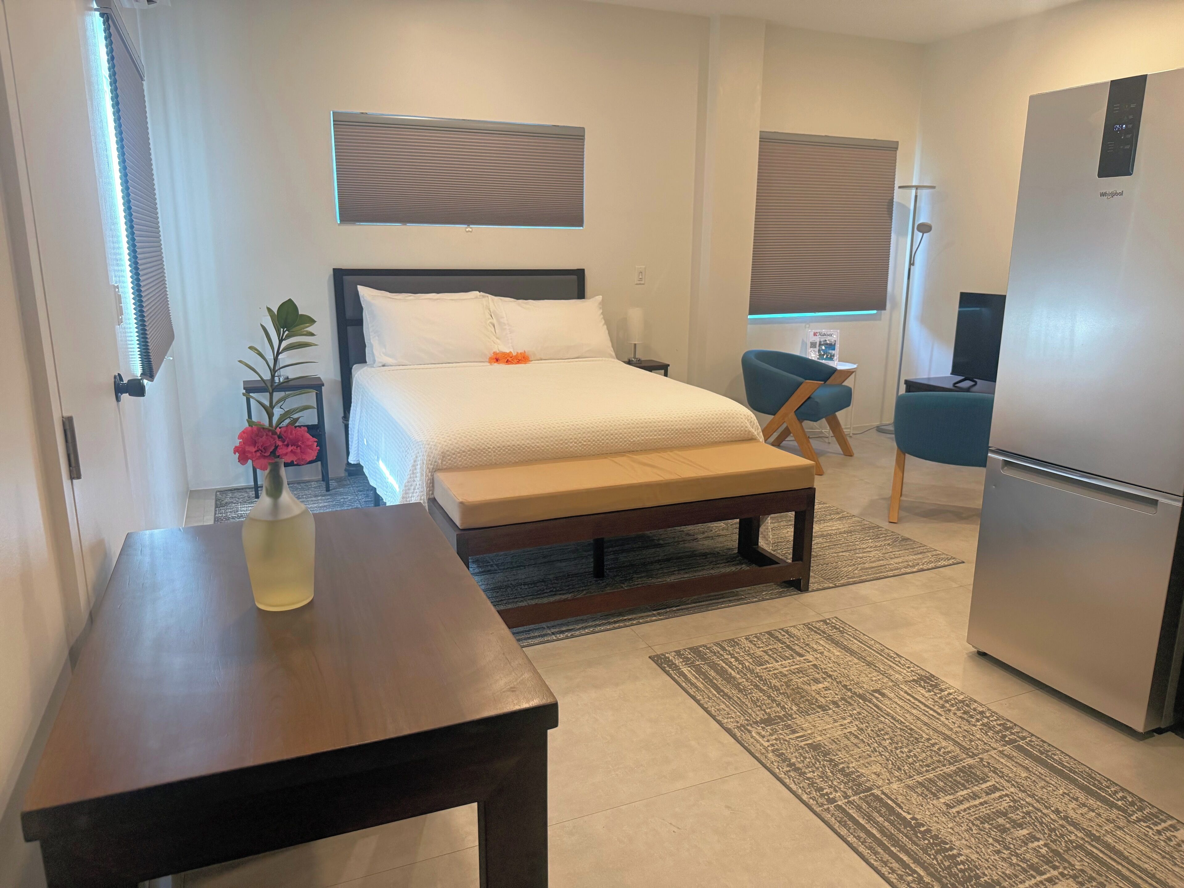 Habitación de hotel con cama, mesita de noche, escritorio pequeño con silla, refrigerador y ventana con persianas.