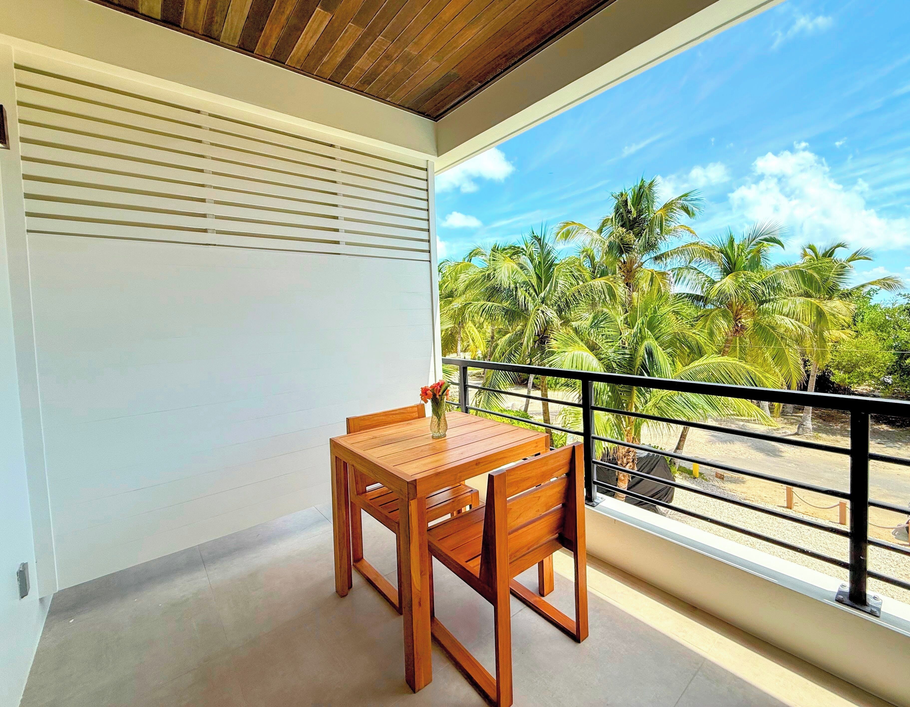 A balcony with a wooden table and chairs, overlooking a beach with palm trees.