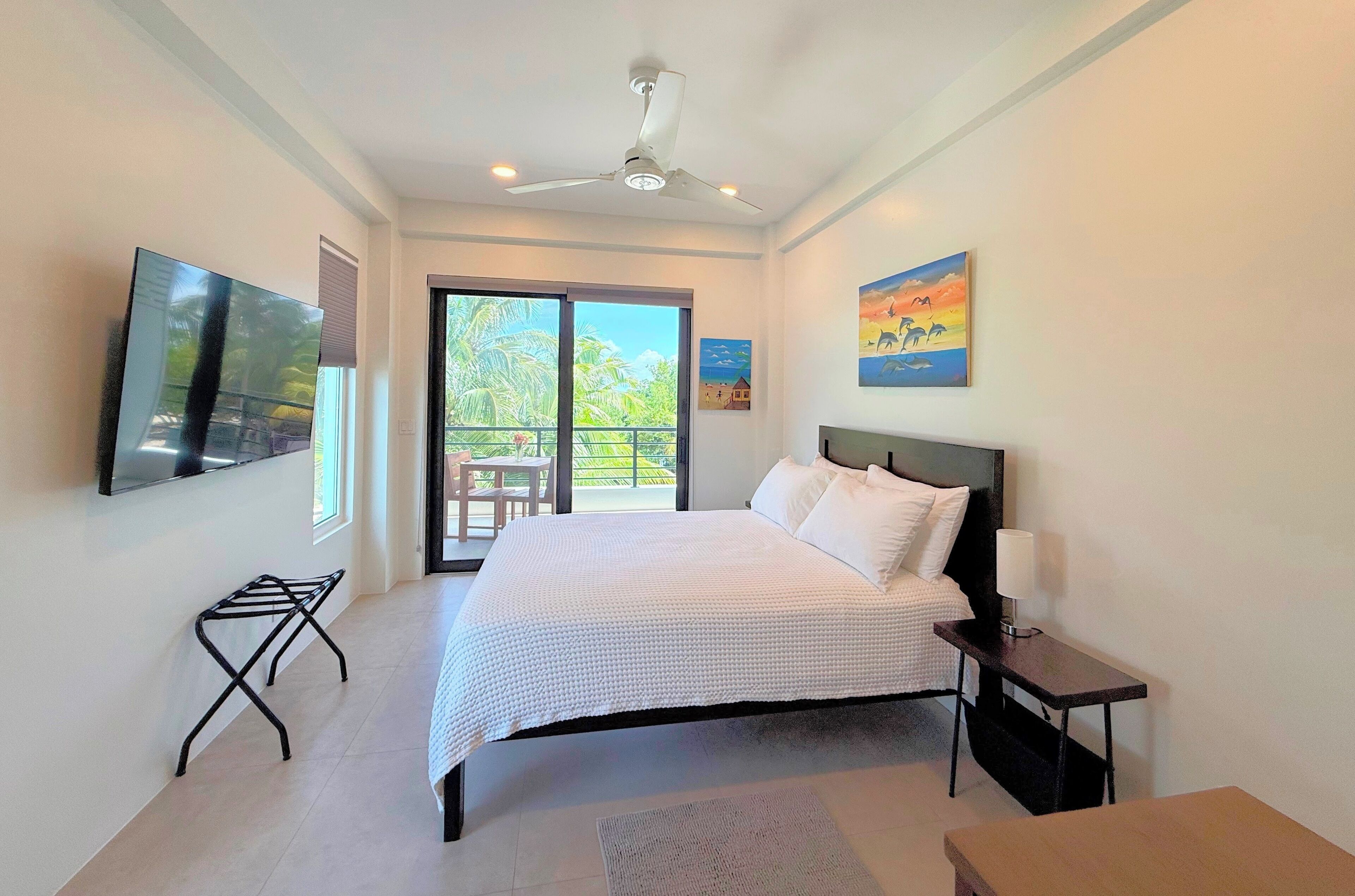 A bedroom with a bed, a ceiling fan, a TV mounted on the wall, and a balcony with a view of palm trees.