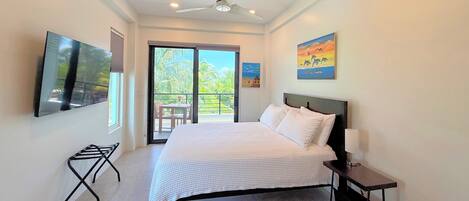A bedroom with a bed, a ceiling fan, a TV mounted on the wall, and a balcony with a view of palm trees.