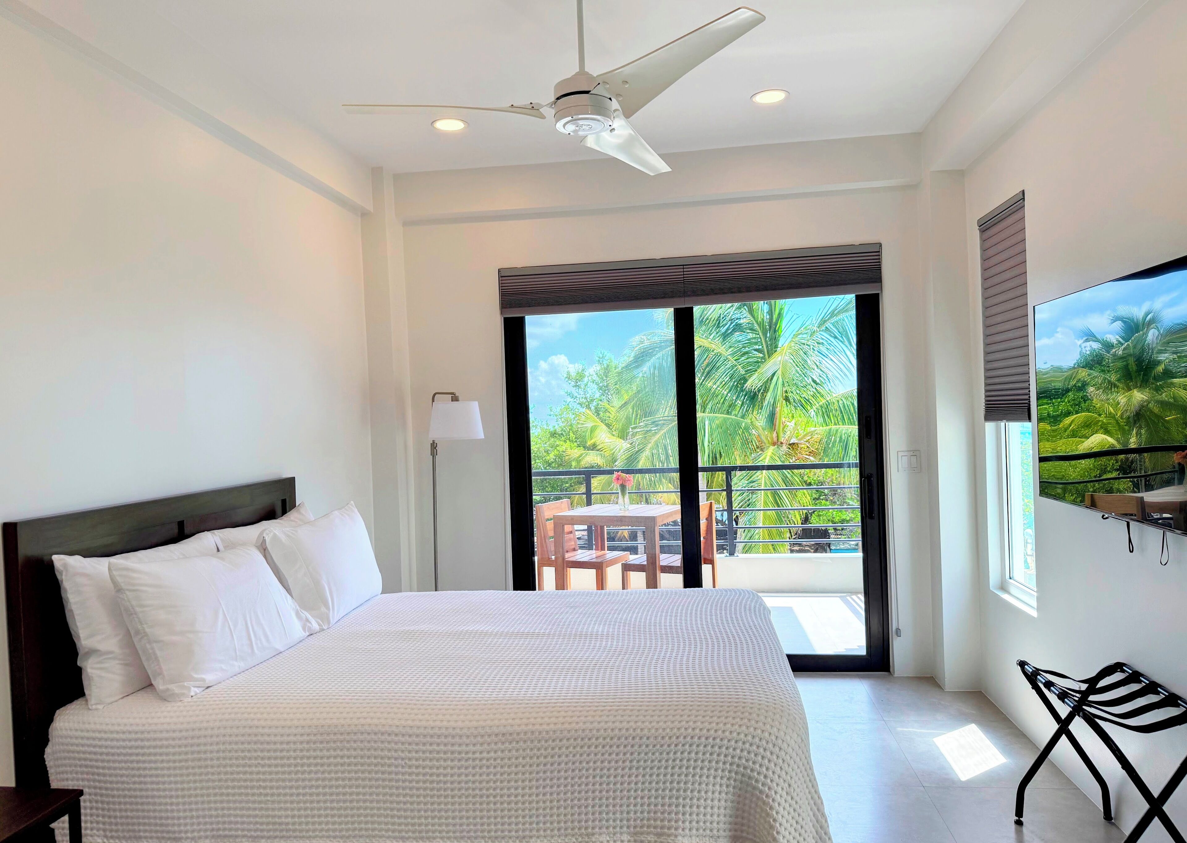 A bedroom with a bed, a ceiling fan, a floor lamp, a balcony with a table and chairs, and a view of palm trees.