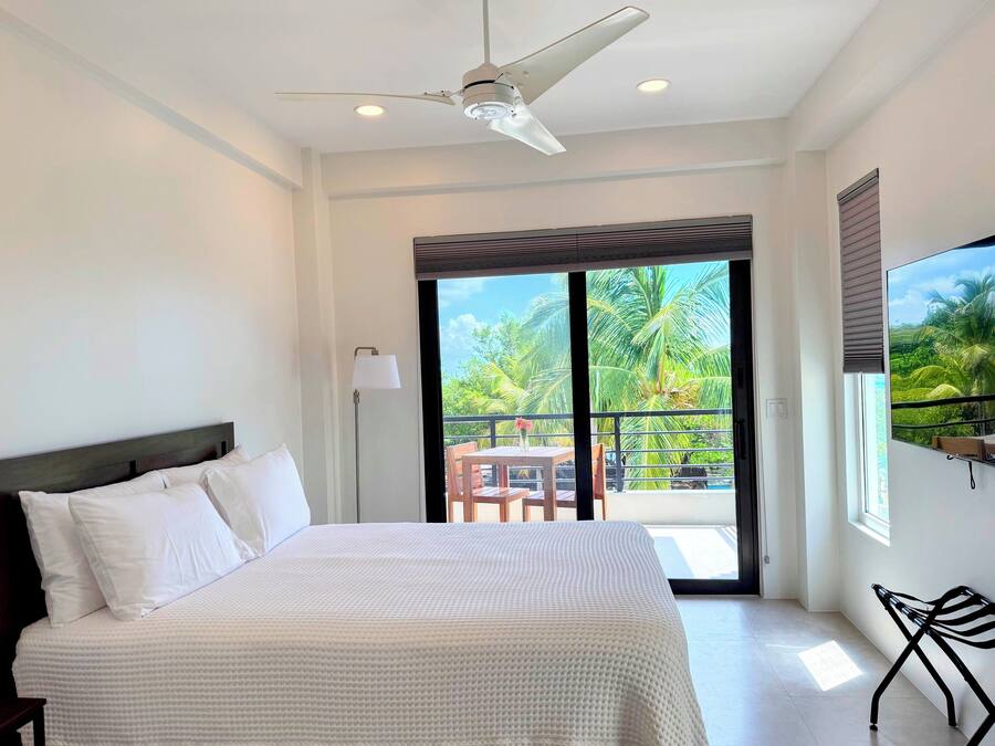 A bedroom with a bed, a ceiling fan, a floor lamp, a balcony with a table and chairs, and a view of palm trees.
