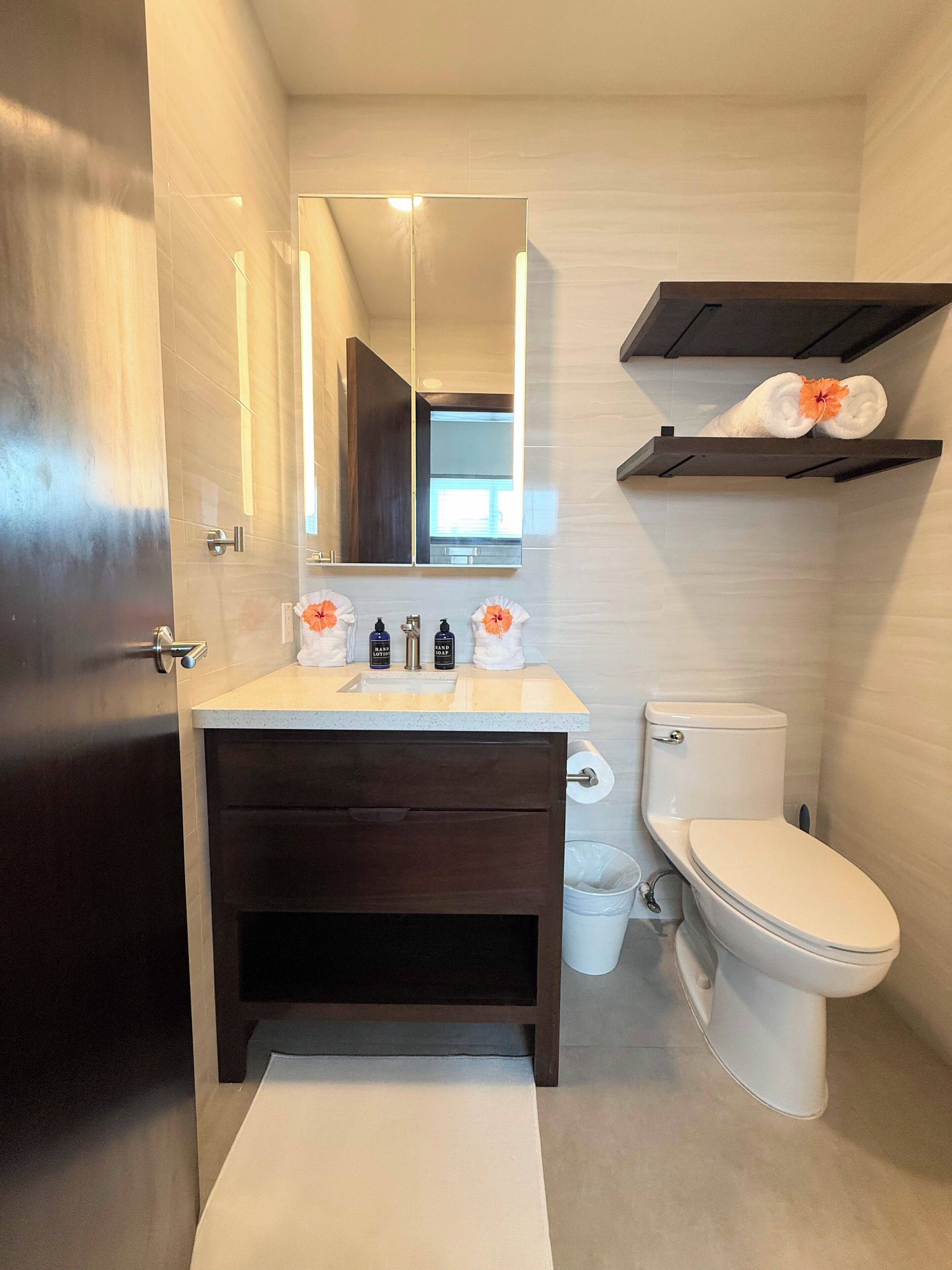 Et moderne badeværelse med et mørkt trækommode, et hvidt toilet, et spejl over vasken og en håndklædestang med en blomst.