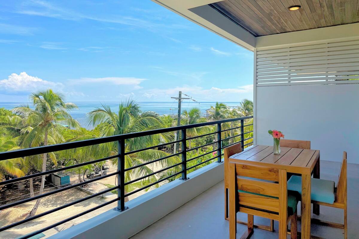 A balcony with a view of the ocean, a wooden table with chairs, and a flower vase.