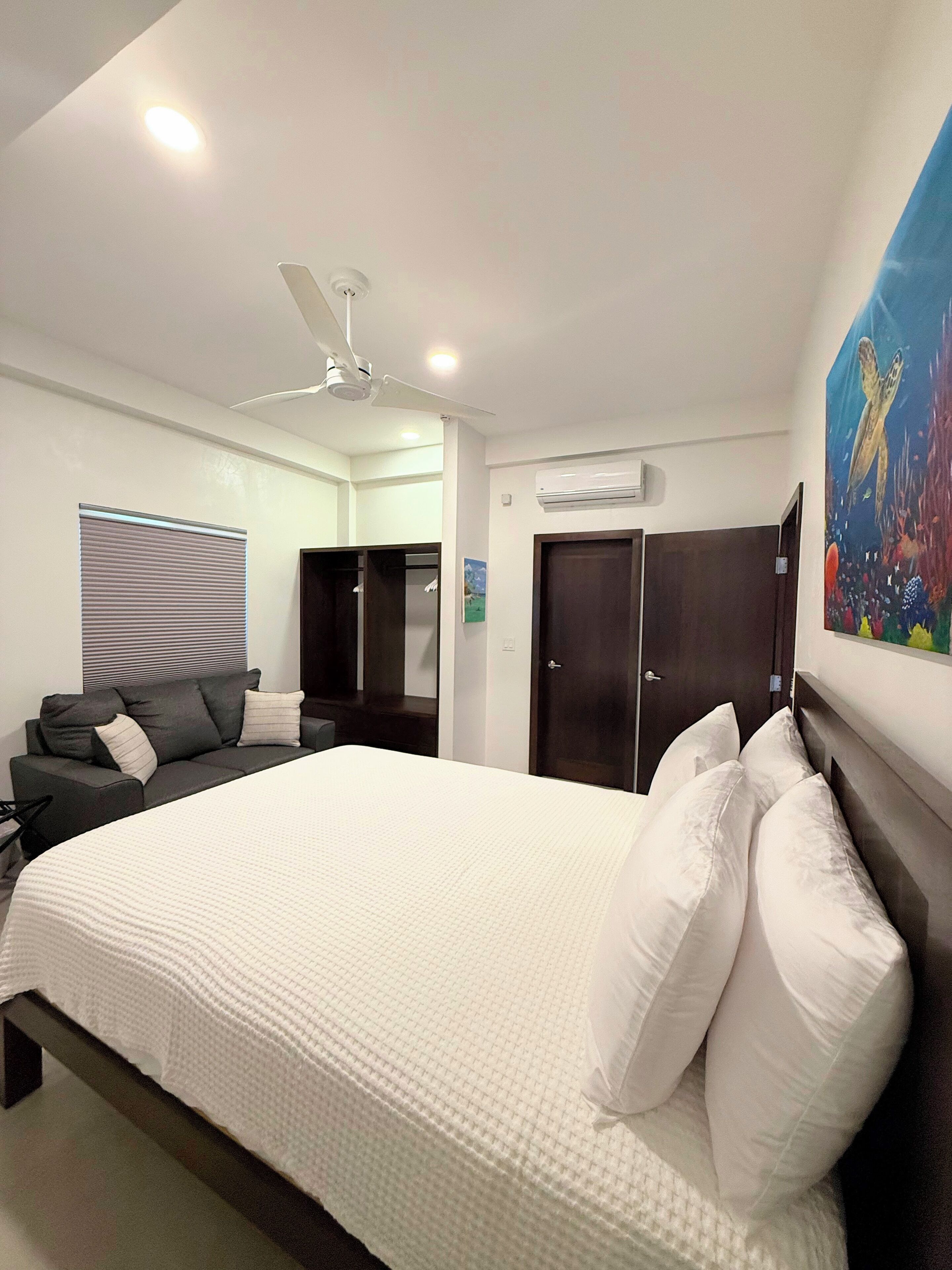 A hotel room with a large bed, a ceiling fan, an air conditioning unit, a dark wooden wardrobe, a dark wooden door, and a painting of a sea turtle on the wall.