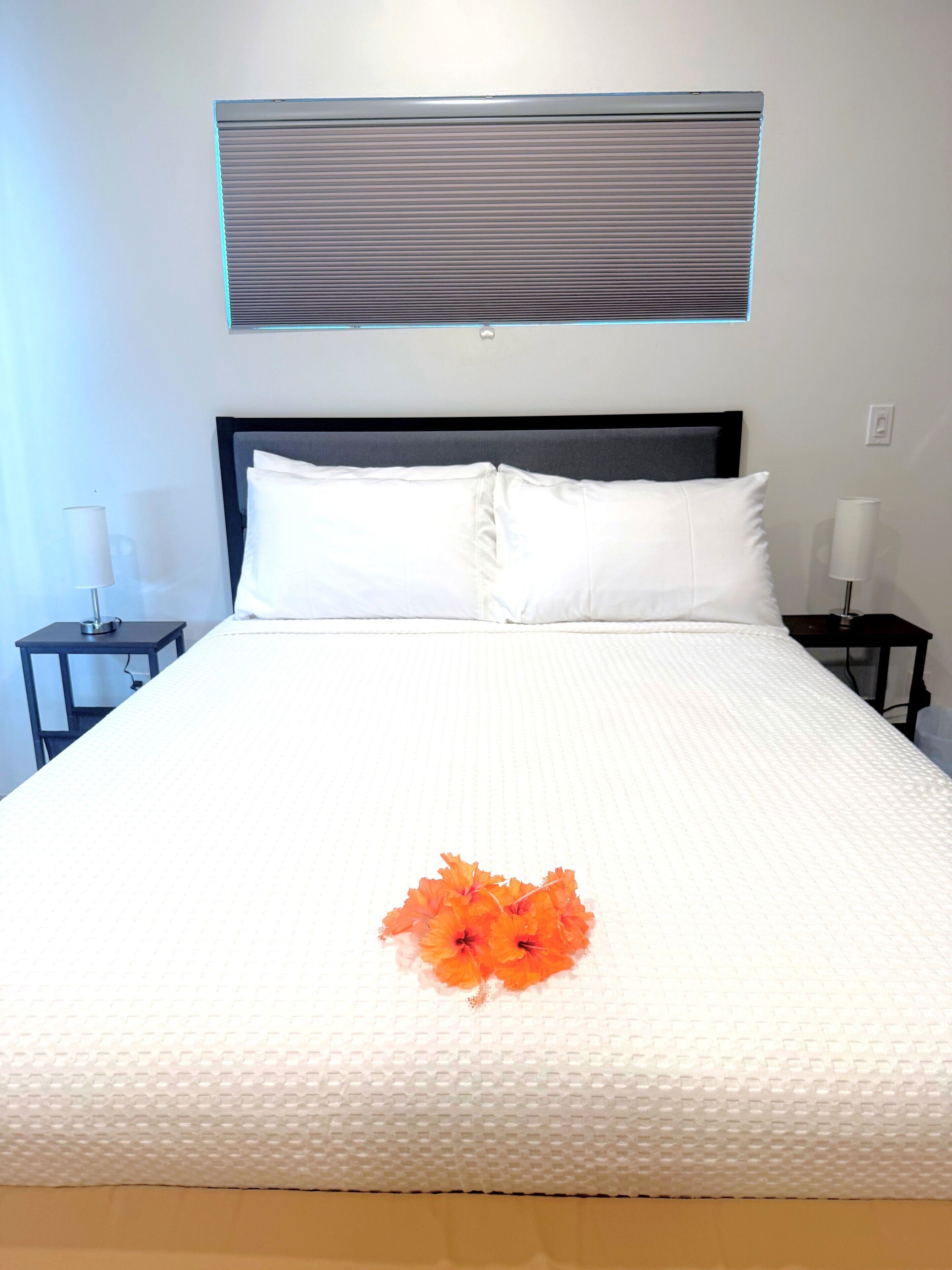 Un lit bien fait, avec du linge de lit blanc, une tête de lit noire et une seule fleur orange posée sur la couverture.