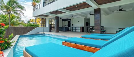 Un área de piscina moderna con sillones reclinables azules y un edificio de dos pisos con balcones.