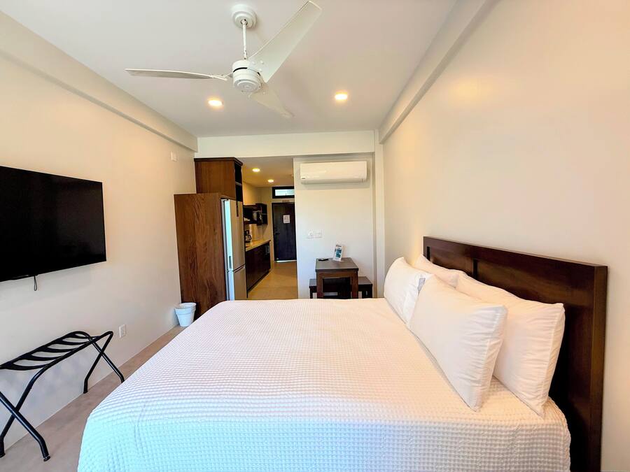 A hotel room with a large bed, a ceiling fan, a flat-screen TV, a kitchenette, and an air conditioning unit.