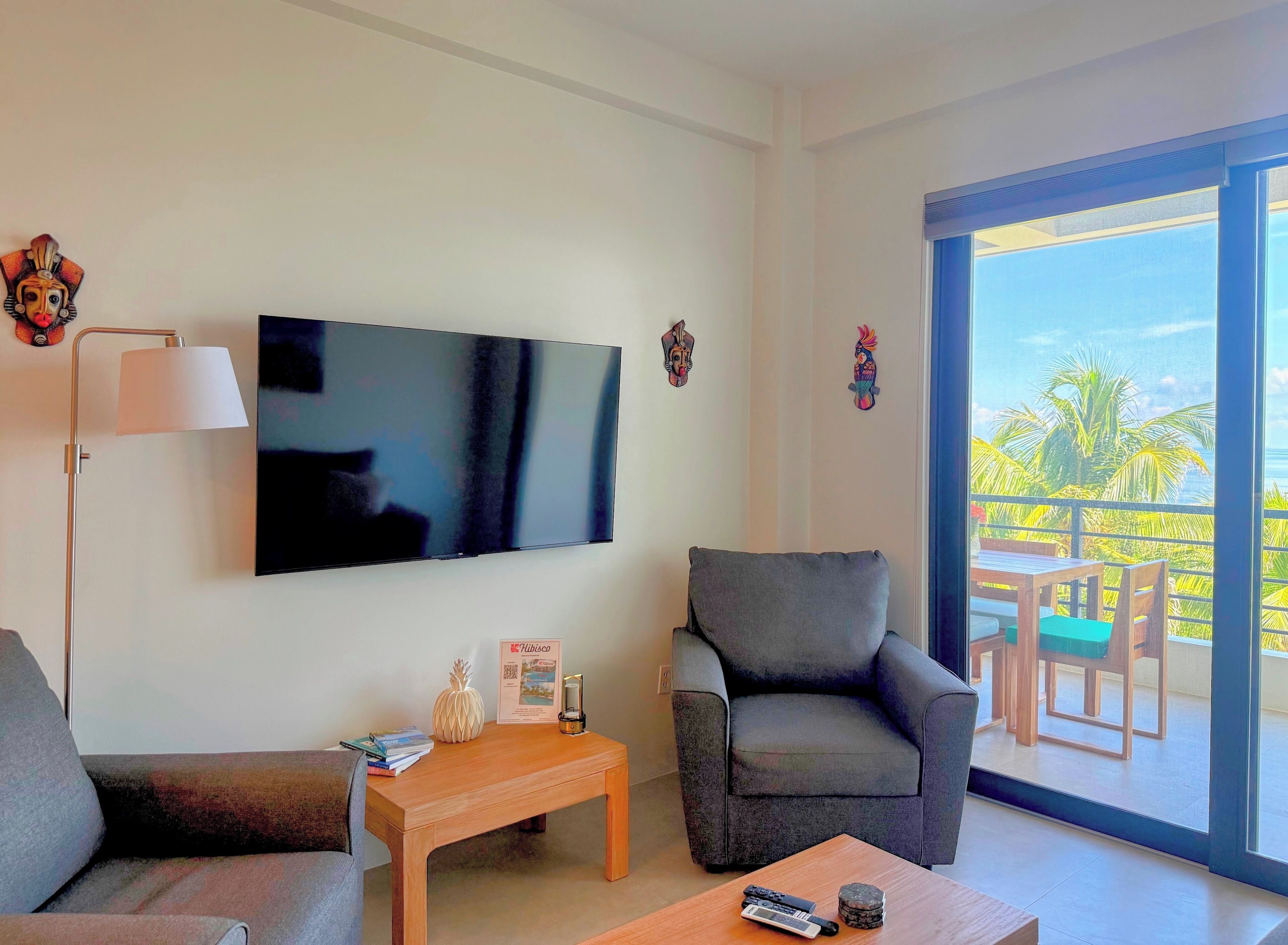 Una sala moderna con televisor de pantalla plana, dos sillones grises, una mesa de centro de madera y una mesita con lámpara y objetos decorativos. Se ve un balcón con muebles de madera y vistas a palmeras.