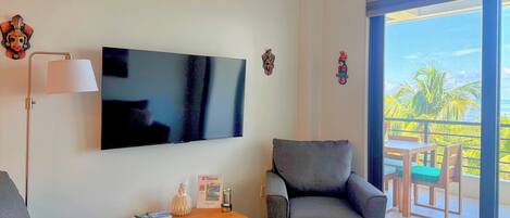 Una sala moderna con televisor de pantalla plana, dos sillones grises, una mesa de centro de madera y una mesita con lámpara y objetos decorativos. Se ve un balcón con muebles de madera y vistas a palmeras.