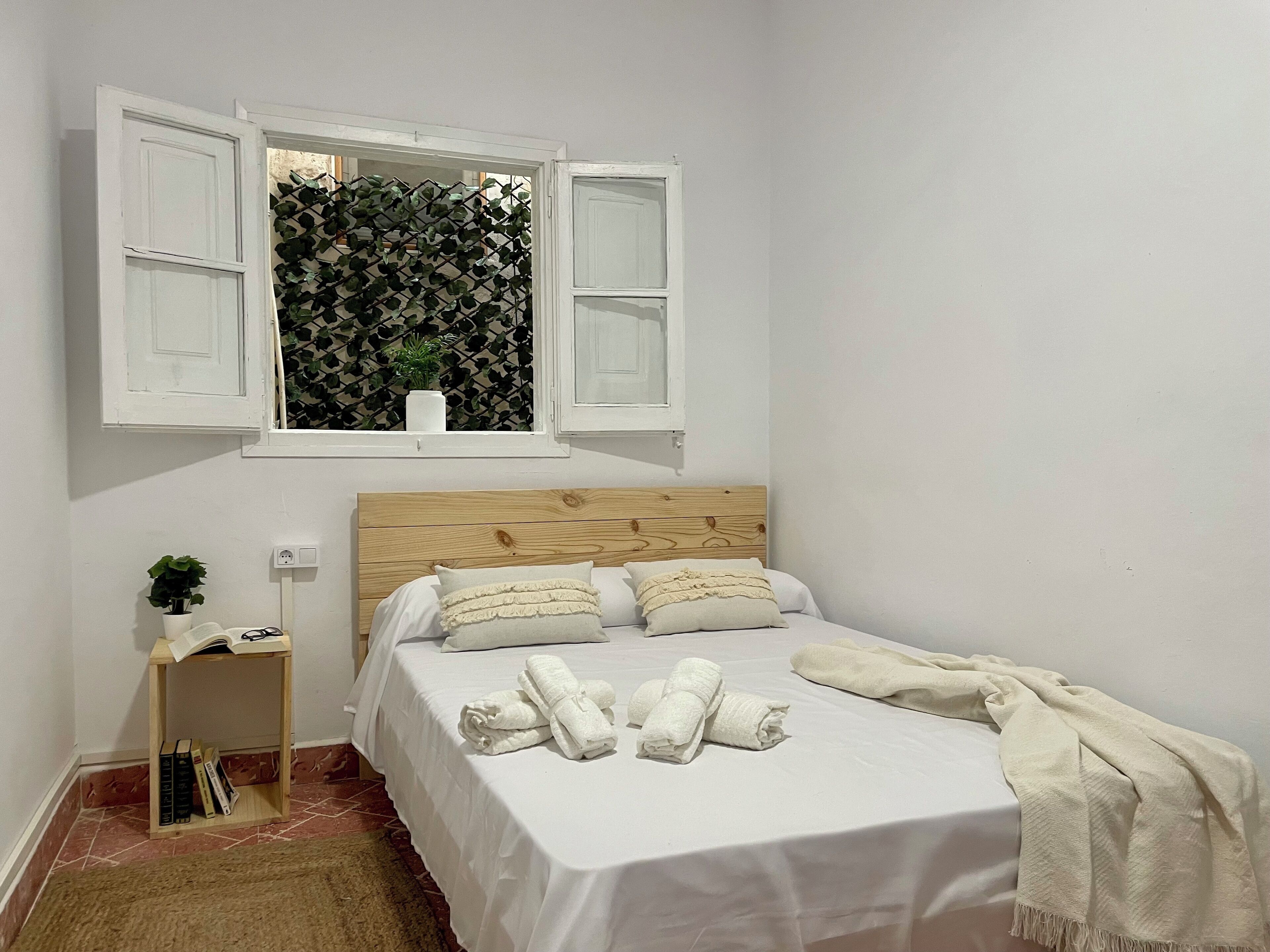 A bedroom with a wooden headboard, a bed with white linens, a window with open shutters, a small bedside table with a plant, and a rug on the floor.