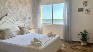 A hotel room with a large bed, a view of the sea, a potted plant, and a patterned wallpaper.