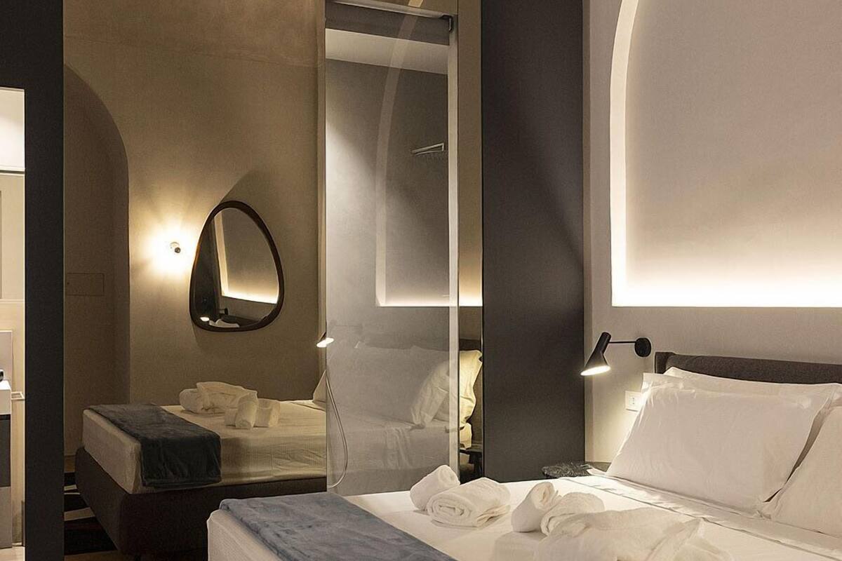 Quarto de hotel moderno com cama grande, mesa de cabeceira, espelho e luminária fixada na parede.