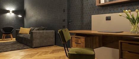 A modern interior with a herringbone wooden floor, a grey sofa, a green chair, a wooden desk, and a wall with a geometric pattern.