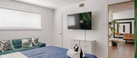 Une chambre à coucher comprenant un lit, un téléviseur, un ventilateur de plafond, une fenêtre avec des stores et une porte donnant sur une autre pièce.