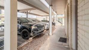A covered parking area with vehicles, a concrete pathway, and a building with a brick facade. - Pink 1BHK, 5 miles from Downtown + Free Parking (Houston)