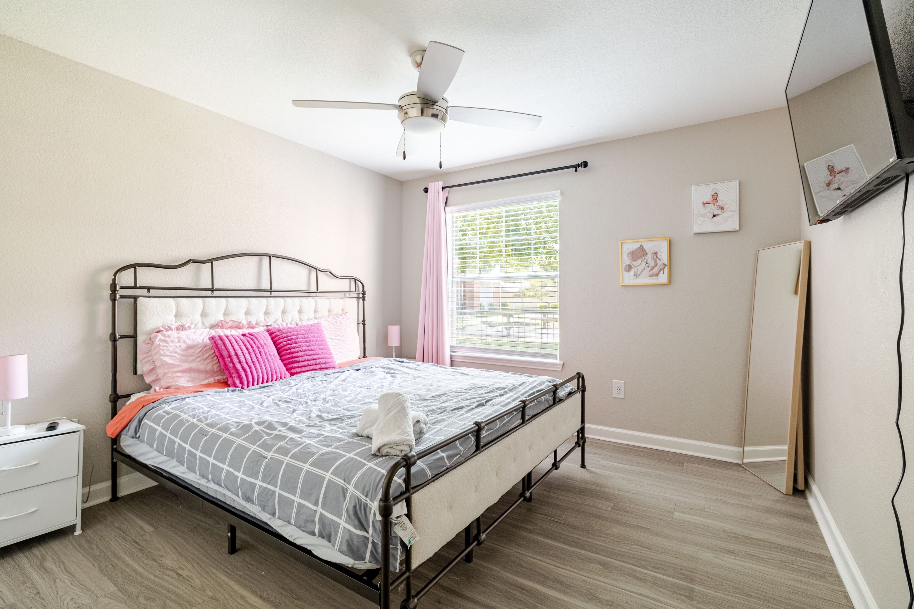 A bedroom with a bed, a nightstand, a bedside lamp, a ceiling fan, a window with curtains, and a wall-mounted TV.
