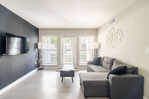 A modern living room with a flat-screen TV, a gray sofa, a coffee table, and a decorative wall piece. - Gothic Blush King 2Bed near Texas Medical Center & in front of NRG Stadium (Houston)