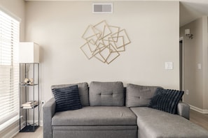 A living room with a sectional sofa, a wall-mounted light fixture, and a radiator. - Gothic Blush King 2Bed near Texas Medical Center & in front of NRG Stadium (Houston)