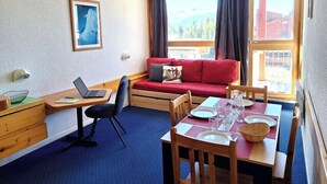 Ein gemütliches Zimmer mit einem roten Sofa, einem für zwei Personen gedeckten Esstisch, einem Laptop auf einem Schreibtisch und einem Bild an der Wand.