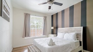 Ein Schlafzimmer mit einem Bett, einem Nachttisch, einer Lampe, einem Deckenventilator, einem Fenster mit Vorhängen und einer Holzbank.