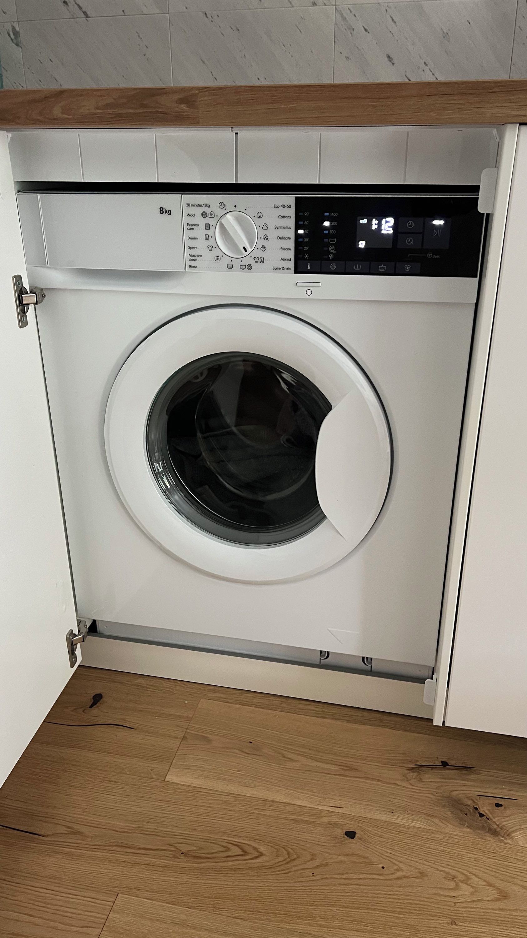 Un lave-linge à chargement frontal doté d’un panneau de commande, situé dans une pièce au plancher en bois et aux murs blancs.