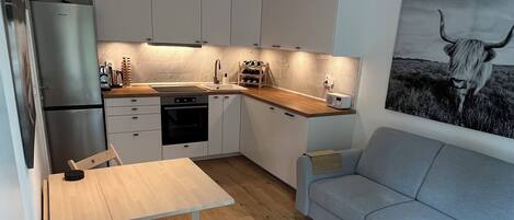 A compact kitchen with white cabinets, a wooden dining table, and a grey sofa.