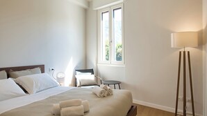 A neatly made bed with rolled towels, a bedside table with a lamp, a chair, and a window with a view of greenery.