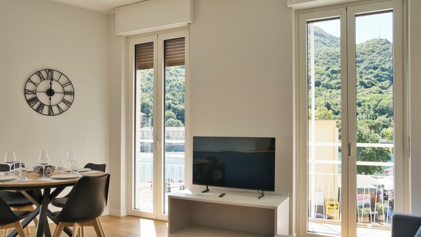 A modern living room with a dining area, a flat-screen TV, and large sliding glass doors leading to a balcony with a view of a green hillside.
