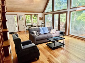 A spacious living room with a wooden ceiling, large windows, a grey sofa, a black armchair, and a dining area with a table and chairs.