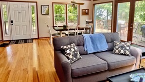 Ein modernes Wohnzimmer mit einem grauen Sofa, gemusterten Kissen und einer blauen Decke. Es gibt einen Essbereich mit Tisch und Stühlen, und große Fenster mit Blick ins Freie.