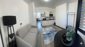 A modern living room with a sofa, a coffee table, and a lamp. There is a kitchen area in the background with a microwave and cabinets.