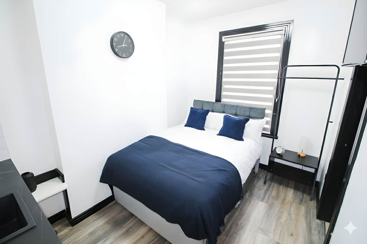 A modern bedroom with a bed, a bedside table, a clock, and a window with blinds.