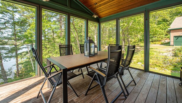 A covered deck with a wooden table and chairs, large windows, and a view of a lake and trees. - Windward Lodge- stunning, dog-friendly private waterfront retreat. Seasonal dock (Woolwich)