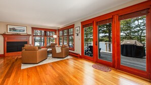 A spacious living room with hardwood flooring, a fireplace, a sofa, and a dining area. Large windows and sliding glass doors provide views of the outdoors. - Windward Lodge- stunning, dog-friendly private waterfront retreat. Seasonal dock (Woolwich)
