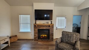 Ein Wohnzimmer mit Kamin, einem darüber hängenden Fernseher und zwei Fenstern mit Jalousien.