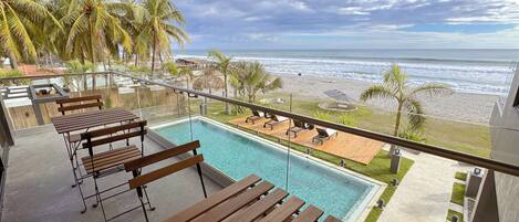Zona de piscina con bancos de madera, vista a la playa y palmeras.