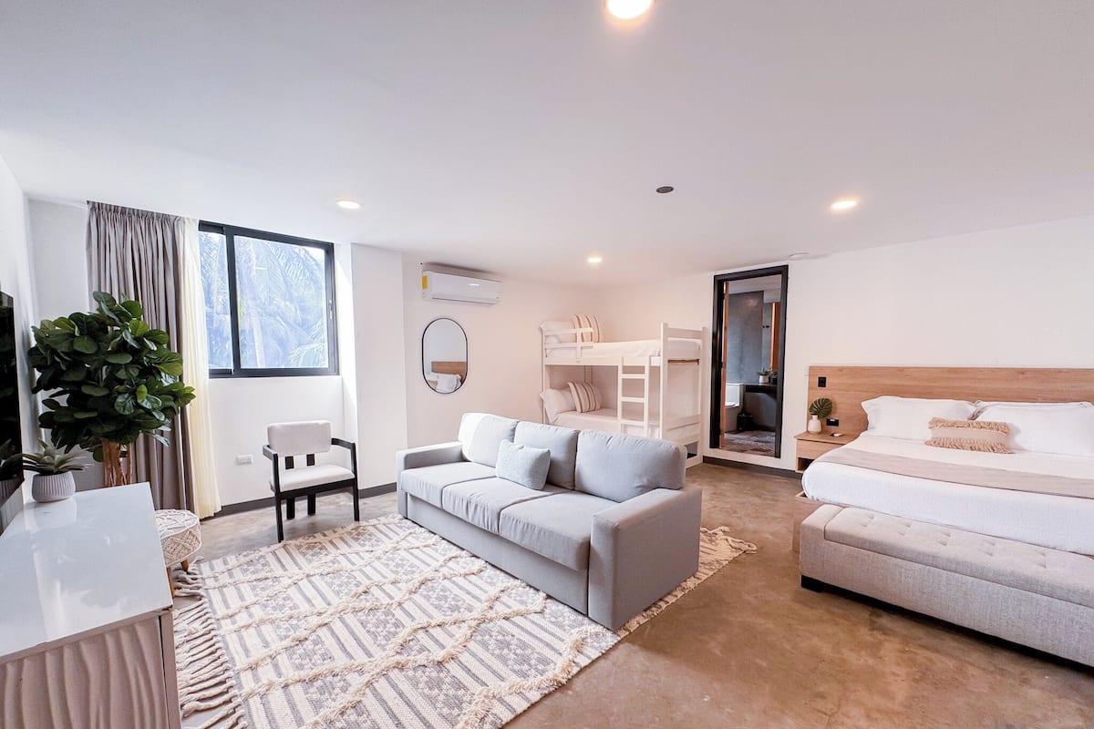 Habitació moderna i ben il·luminada amb un sofà, un llit, una taula de treball i una finestra gran.