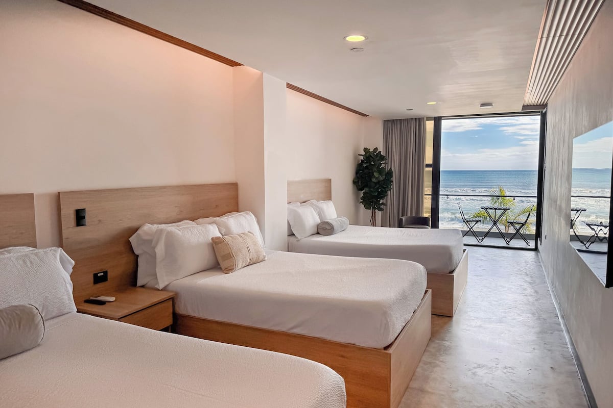 Una habitació d'hotel moderna amb dos llits, una finestra gran amb vista a la platja i un balcó amb mobiliari exterior.