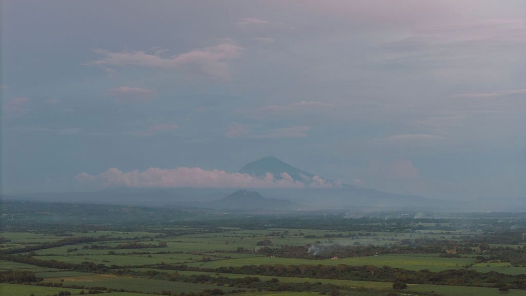 A mountain landscape with a prominent peak, surrounded by green fields and a few scattered buildings.