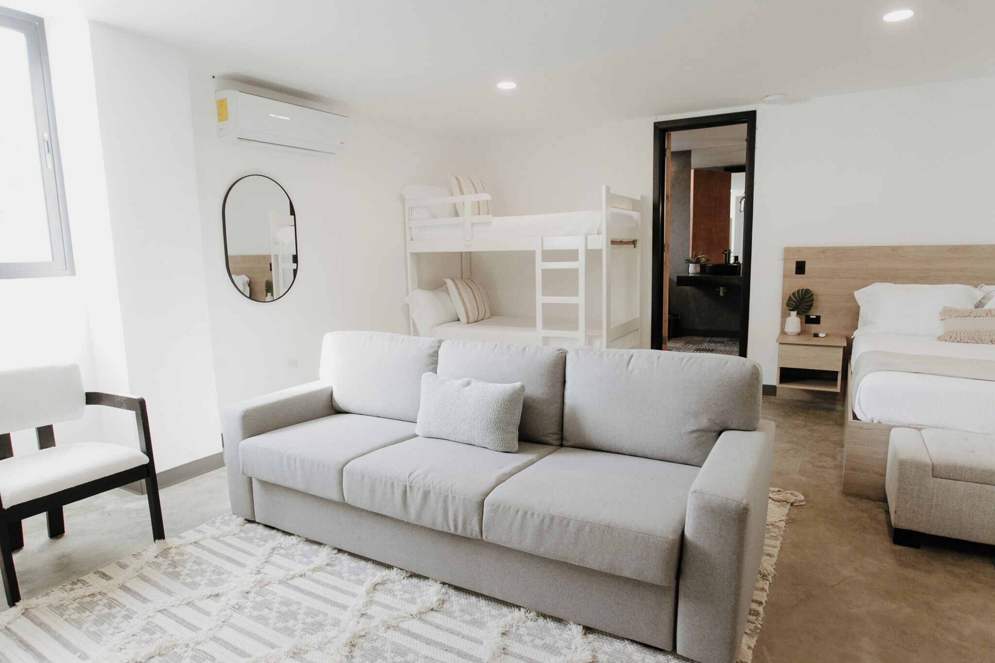 A modern, well-lit room with a grey sofa, a bunk bed, a wooden headboard bed, a mirror, and a small plant.