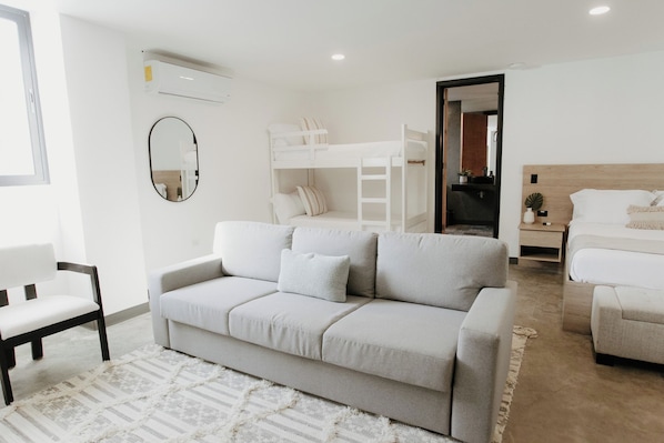 Ein modernes, helles Zimmer mit einem grauen Sofa, einem Etagenbett, einem Bett mit Holz-Kopfteil, einem Spiegel und einer kleinen Pflanze.