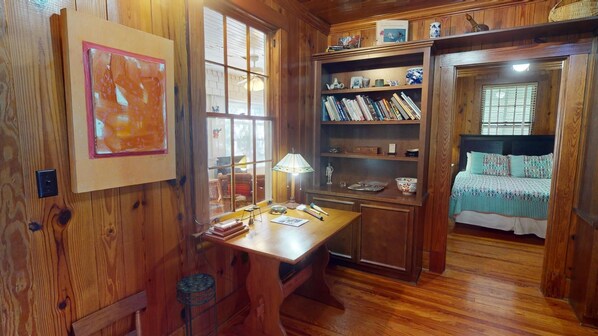 A cozy room with wooden walls and floors, a desk with a lamp, a bookshelf, and a bed with patterned bedding.