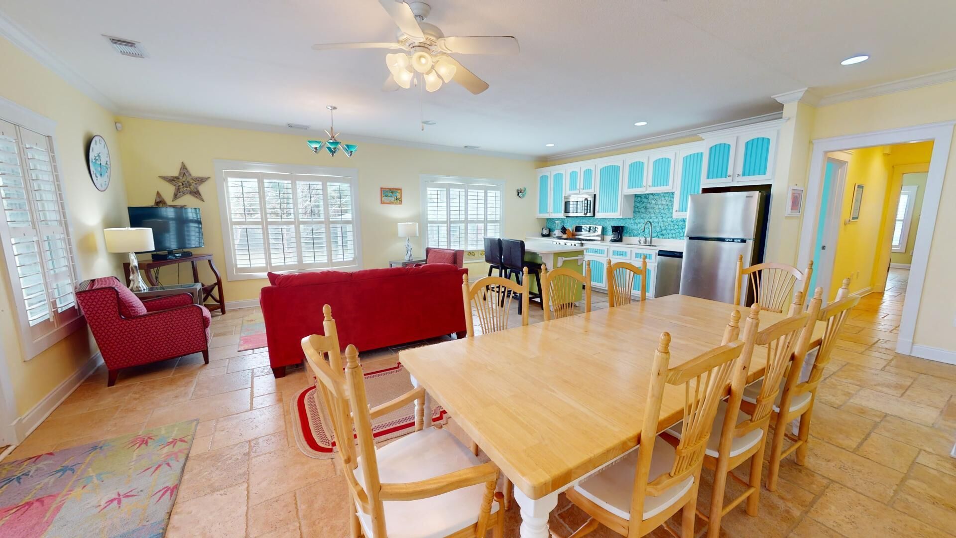 A spacious living room with a red sofa, a wooden dining table with chairs, a kitchen with blue cabinets, and a ceiling fan.