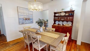 Un coin repas avec une table en bois, des chaises, un lustre, une bibliothèque où sont exposées des assiettes et un tableau accroché au mur.